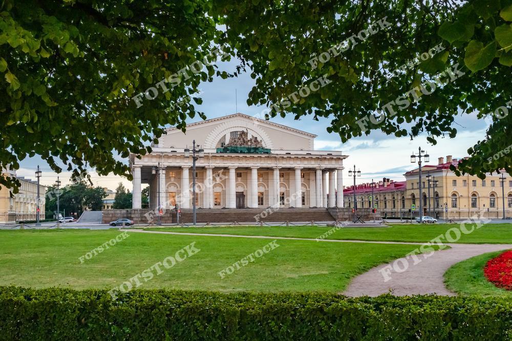 На фотографии изображено здание Биржи, расположенное на Стрелке Васильевского острова в Санкт-Петербурге. Построенное в стиле классицизма, оно является ярким образцом архитектуры XIX века. На переднем плане зеленый газон и раскидистые деревья, которые добавляют летней свежести композиции. Это место – одна из главных достопримечательностей города, часто посещаемая туристами.